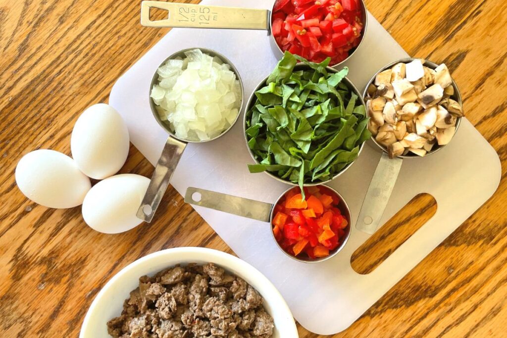 Onion, tomatoes, spinach, mushrooms, sausage, bell peppers, eggs on a cutting board