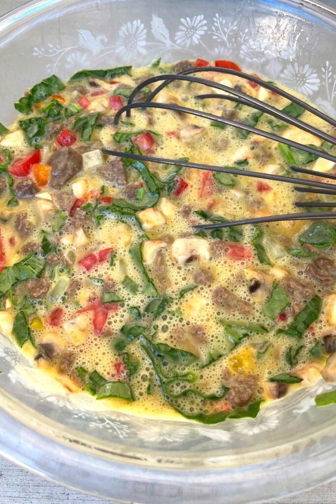 raw scrambled with vegetables and sausage in glass mixing bowl with whisk